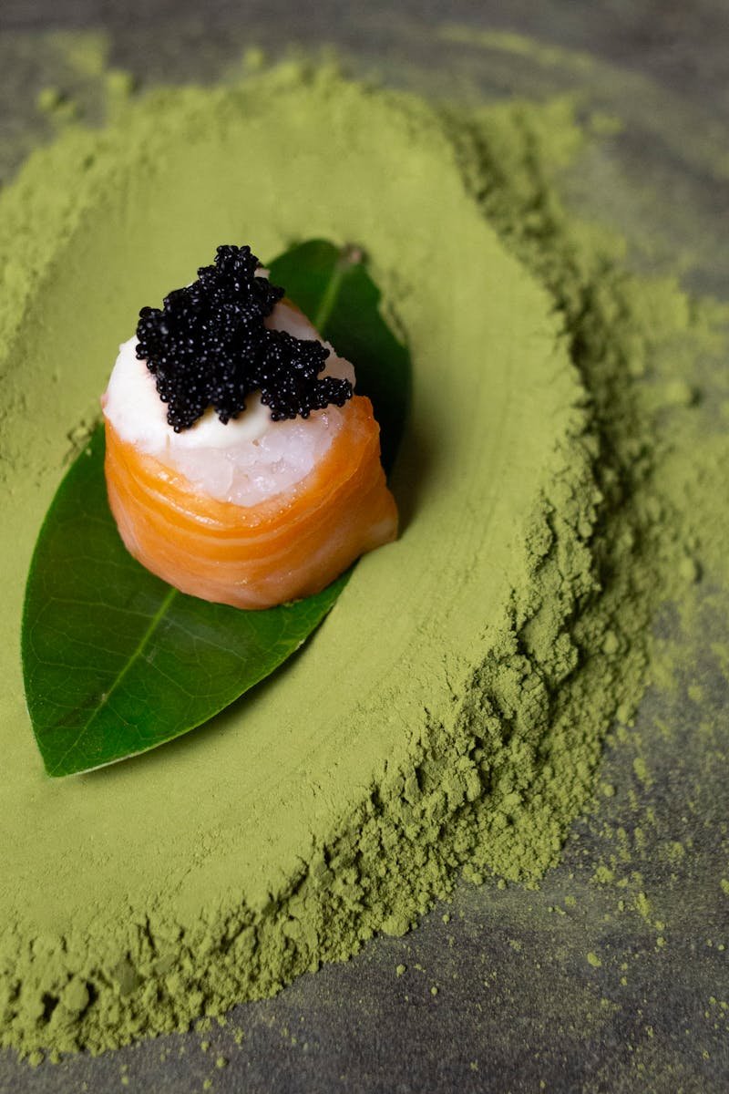 A close-up of salmon sushi topped with fish roe, placed on a leaf surrounded by matcha powder.